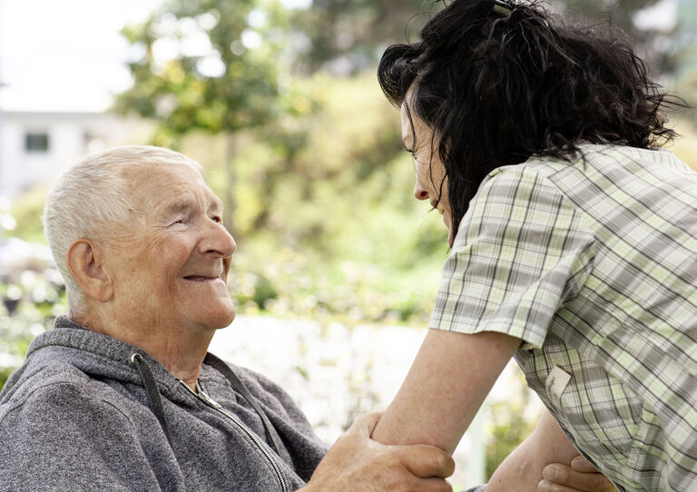 Mitarbeiterin lehnt sich zu Bewohner im Rollstuhl, er hält sie an den Händen Mitarbeiterin lehnt sich zu Bewohner im Rollstuhl, er hält sie an den Händen