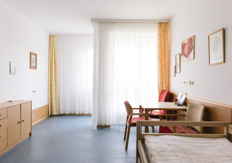 Ein Zimmer im Clementinum mit großen Fenstern, hellblauem Boden, Bett, Tisch und Stühlen sowie einer Kommode.