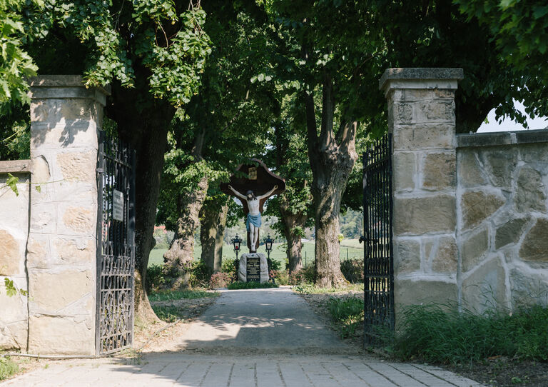 Ein geöffnetes eisernes Tor des Clementinums, das den Blick auf Laubbäume und eine Jesusfigur auf dem Kreuz freigibt.