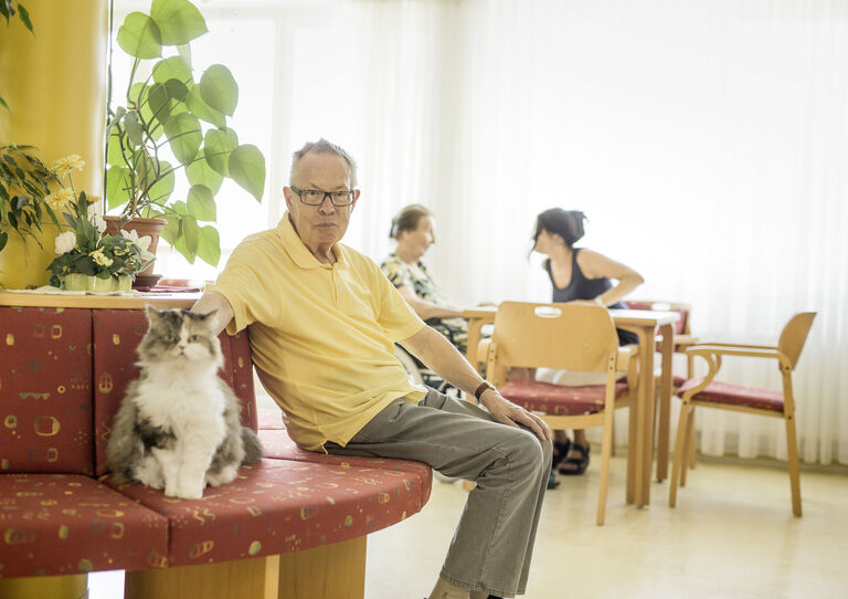 Ein Bewohner streichelt im Wohnbereich eine Katze, die neben ihm auf der Bank sitzt.
