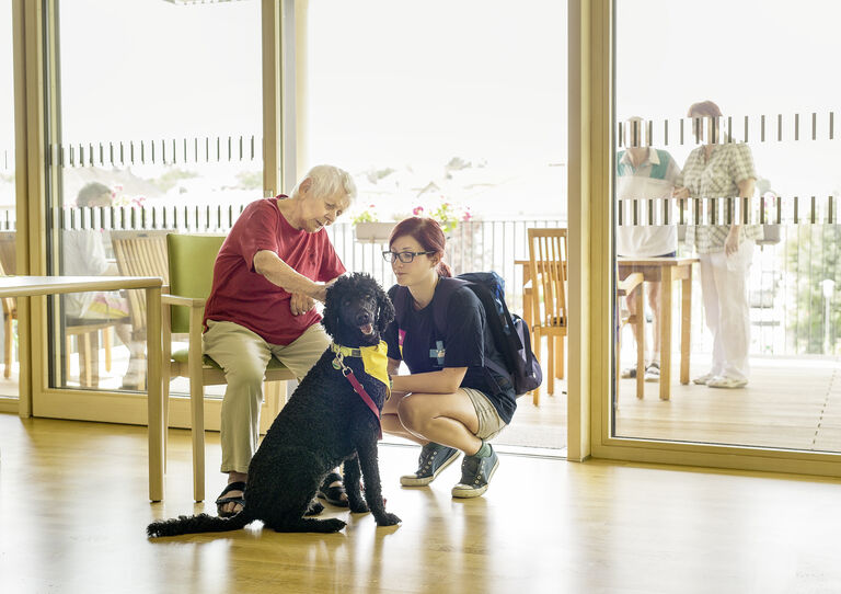 Eine Bewohnerin streichelt einen Therapiehund, die Tiertherapeutin sitzt neben dem Hund.