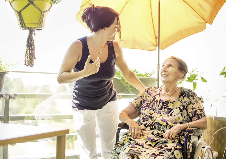 Eine Pflegerin und eine Bewohnerin unterhalten sich auf der Terrasse unter einem Sonnenschirm.