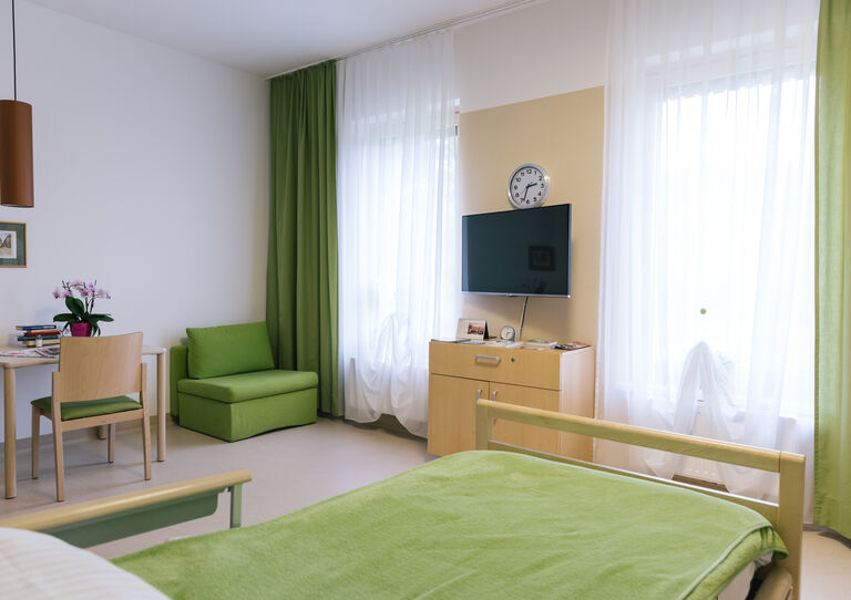Ein Zimmer des Stephansheims mit großen Fenstern, einem Bett mit gründer Tagesdecke, grünen Gardinen und grünem Sessel sowie einem Tisch mit Stuhl und einem Fernseher.
